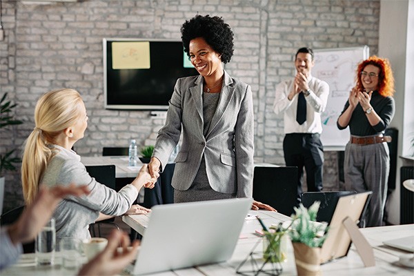 Two business associates shake hands as others applaud in background